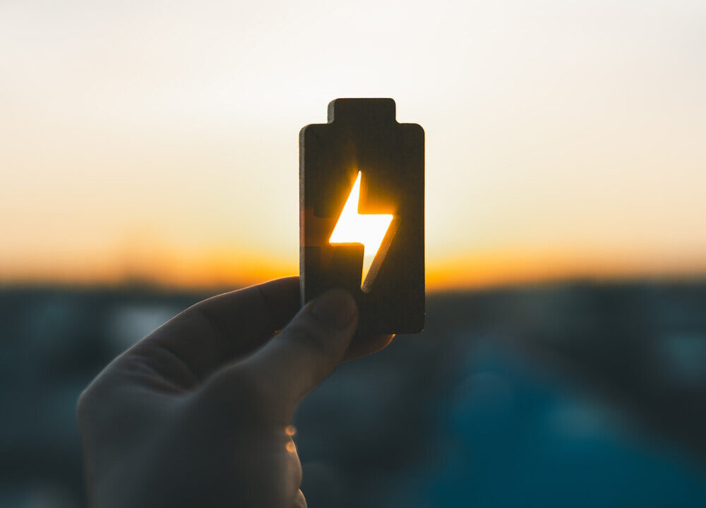 Hand som håller en batteriformad symbol med en blixt i mitten, genom vilken solljus strömmar. Bakgrunden präglas av en varm solnedgång, vilket symboliserar hållbar energi och lagring av förnybar kraft. Bilden fångar essensen av energiomställning och innovation inom grön teknik.